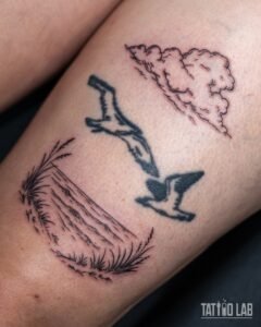 Fine line tattoo of two black seagulls flying between a cloud and a beach dune with tall grass on the thigh