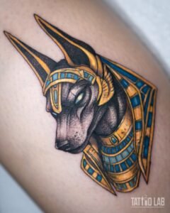 Anubis head tattoo in neo-traditional colour with a gold and turquoise headdress and collar on the leg