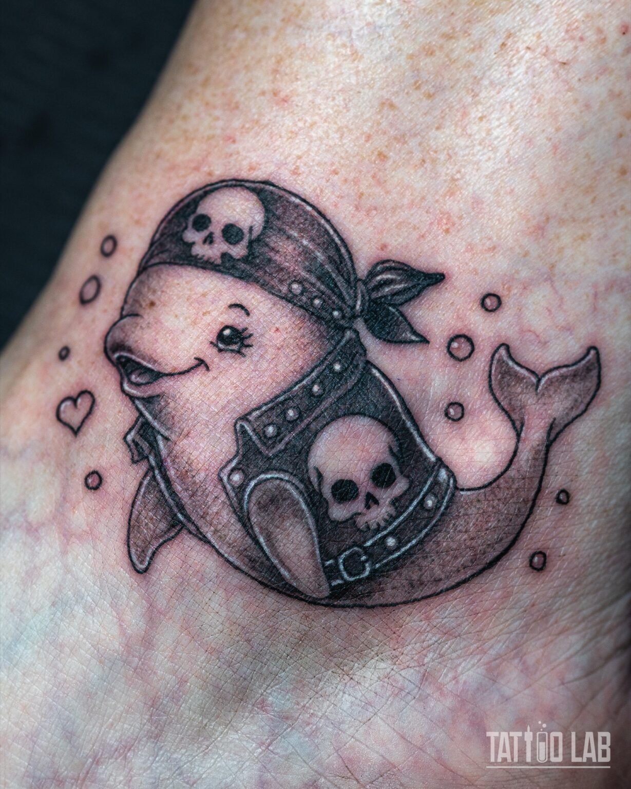 Cartoon beluga-style whale tattoo wearing a skull bandana and studded harness, black and grey, on the foot, Tattoo Lab, Halifax