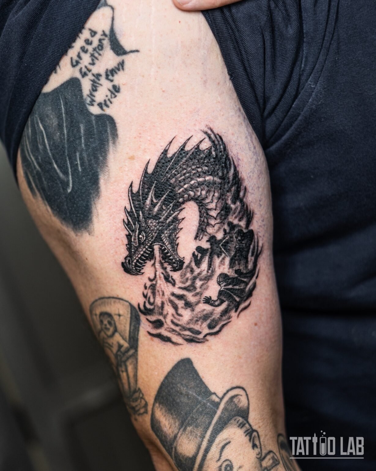 Blackwork dragon tattoo breathing fire over small sword-wielding figures on the upper arm, Tattoo Lab, Halifax