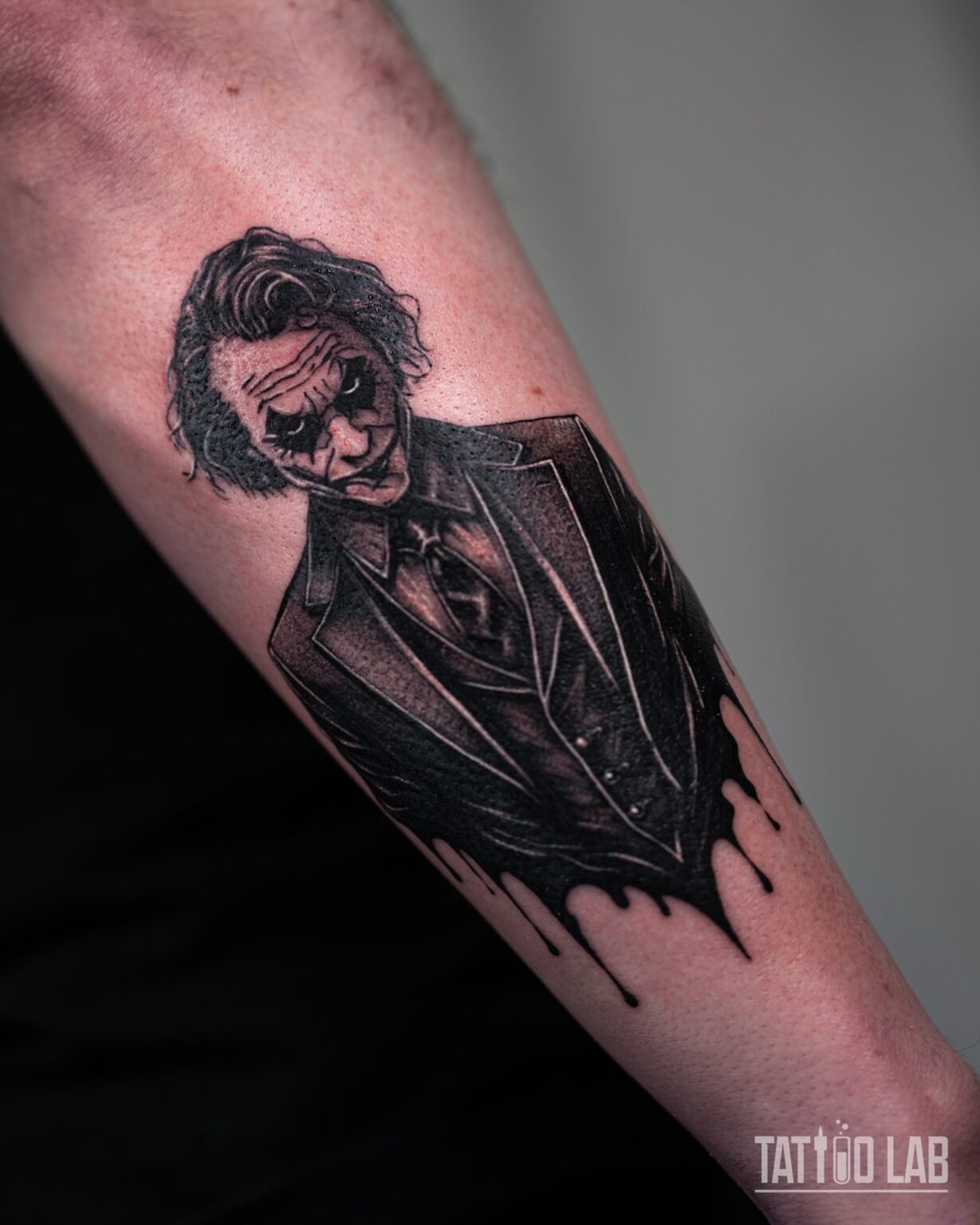 Black and grey Joker-inspired villain portrait tattoo in a suit with black ink drips on the outer forearm, Tattoo Lab, Halifax