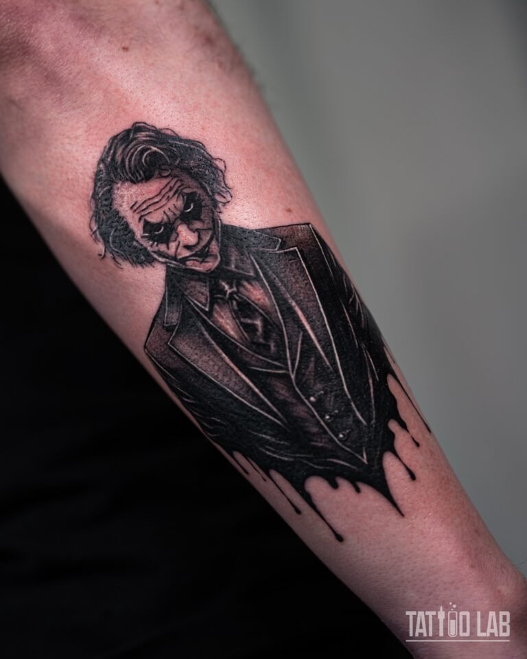 Black and grey Joker-inspired villain portrait tattoo in a suit with black ink drips on the outer forearm