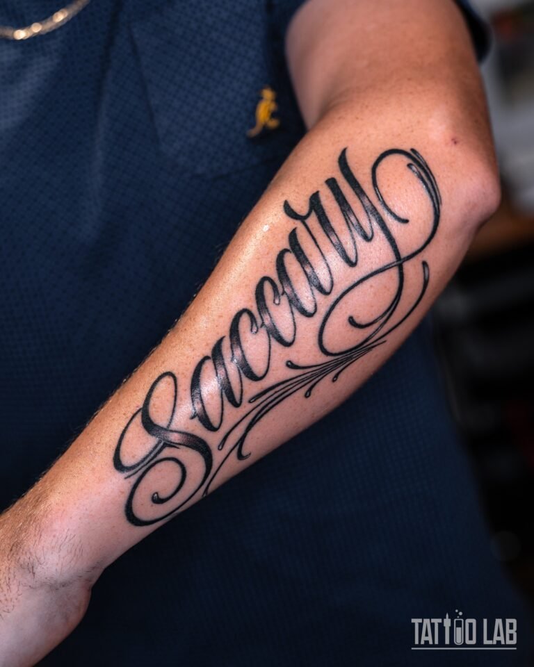Black script lettering tattoo with ornate flourishes running along the forearm